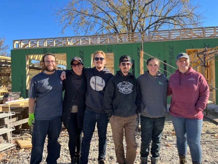 Six smiling people on a Habitat for Humanity construction site standing in front of a house