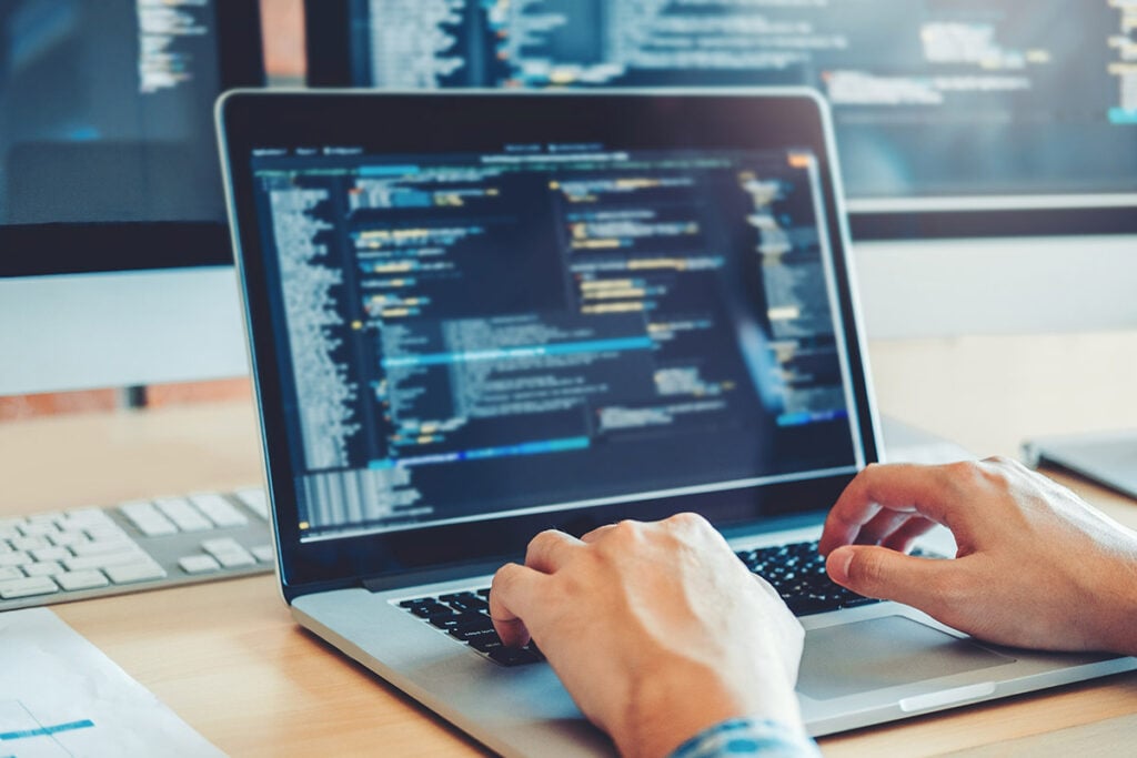 Programmer working on a website; close up shot of hands on a laptop wiht two computer screens in the background